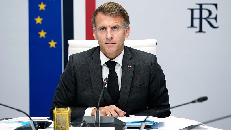 Emmanuel Macron sentado en una sala institucional ante una mesa con documentos, vistiendo traje formal y luciendo un distintivo oficial en la solapa.