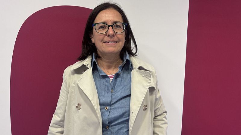 Plano medio de la periodista Teresa Turiera-Puigbò sonriendo, con gabardina beige, camisa vaquera y fondo blanco con franjas granates.