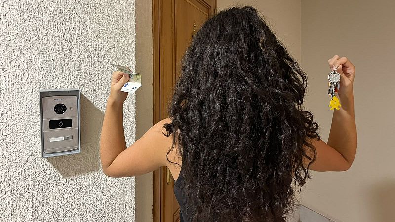 Una mujer de espaldas en un vestíbulo muestra unas llaves y dinero en efectivo, con un videoportero en la pared y una puerta de madera detrás.
