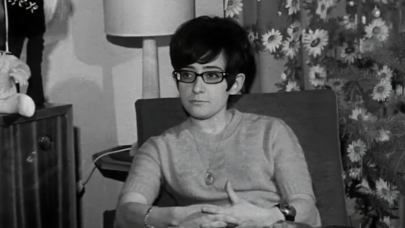 Fotografía en blanco y negro de la joven música Maryní Callejo sentada en una butaca, con gafas oscuras, jersey de punto y una expresión serena en un entorno doméstico.