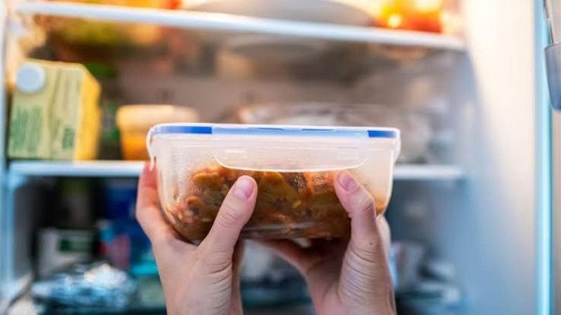 Primer plano de unas manos sosteniendo un t�per con un guiso de verduras ante un frigor�fico abierto.