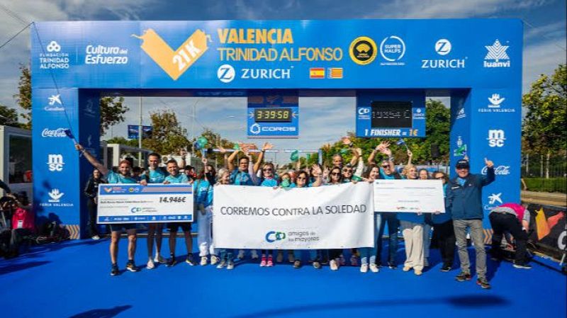 Grupo de personas sonrientes celebrando junto a un arco de meta azul, sosteniendo una pancarta solidaria y un cheque de donación en una carrera en Valencia.