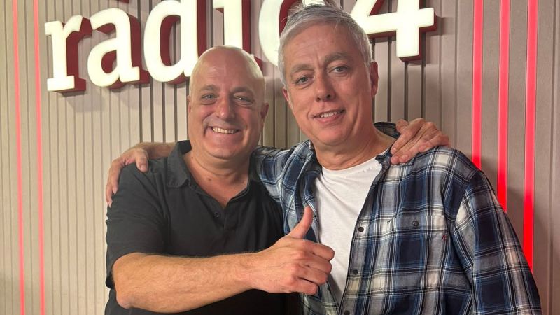 Dos hombres adultos sonrientes posan abrazados en un estudio de radio decorado con paneles de madera y luces de ne�n rojas.