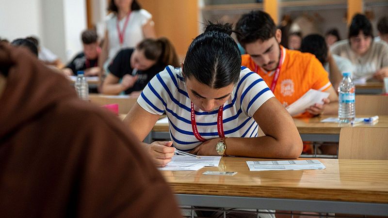 Estudiante joven concentrada redactando su examen en un aula universitaria con fichas identificativas y lanyard rojo visible.