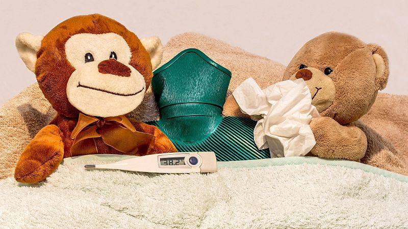 Dos peluches arropados en la cama con un termómetro que marca fiebre de 38,2 °C y una bolsa de agua caliente, representando un resfriado infantil.