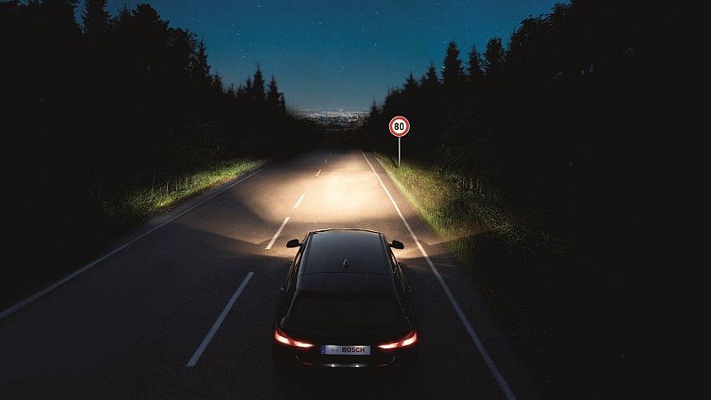 Un coche oscuro circula de noche por una carretera iluminada por sus faros, con una señal de velocidad en el arcén y la silueta de una ciudad al fondo.