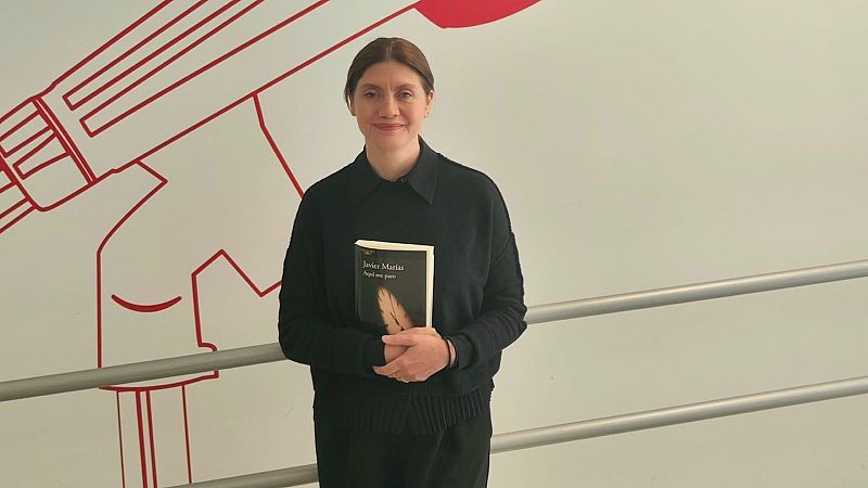La editora Pilar Reyes posa sonriente ante una pared blanca con líneas rojas, sosteniendo el libro póstumo "Aquí me paro" de Javier Marías.