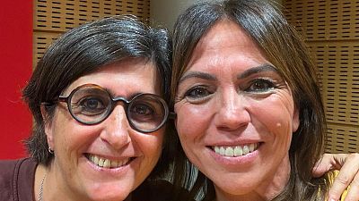Primer plano de dos mujeres sonrientes en un estudio de radio, con paneles ac�sticos de madera clara y una pared roja al fondo.