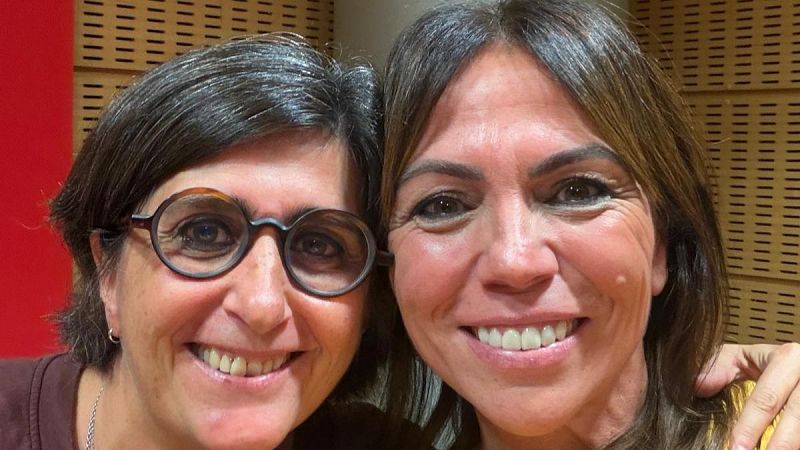 Primer plano de dos mujeres sonrientes en un estudio de radio, con paneles acústicos de madera clara y una pared roja al fondo.