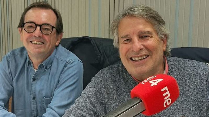 Dos hombres sonrientes en un estudio de radio, con un micrófono con esponja roja en primer plano y paneles acústicos al fondo.