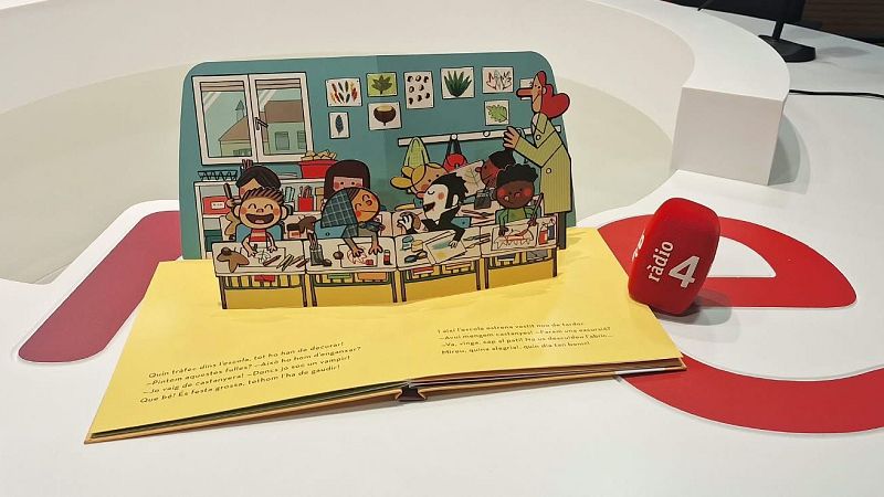 Un libro infantil desplegable con una escena escolar de otoño se encuentra abierto junto a un micrófono rojo de Ràdio 4 sobre una mesa blanca.