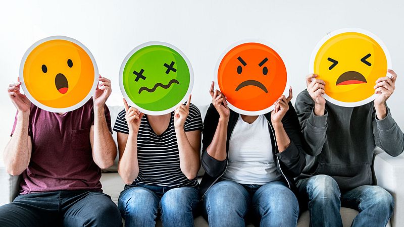Cuatro personas sentadas en un sofá sostienen carteles con emojis que ocultan sus rostros y muestran diversas expresiones emocionales.