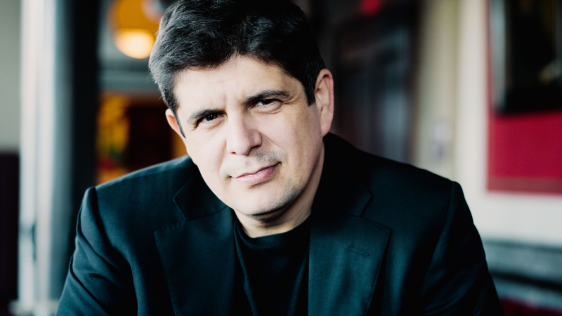 Plano medio del pianista Javier Perianes mirando de frente con expresi�n serena, vistiendo ropa oscura y elegante ante un fondo difuminado con luces c�lidas.