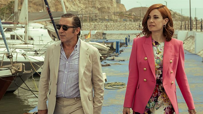 Un hombre con traje beige y gafas de sol camina junto a una mujer con americana fucsia por el espigón de un puerto deportivo con barcos al fondo.