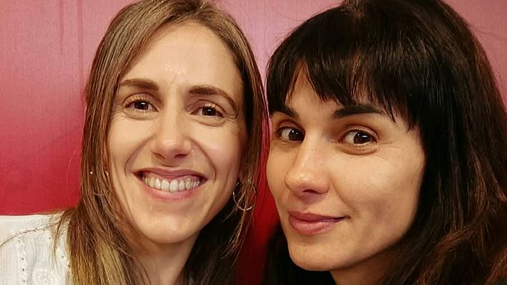 Primer plano de dos mujeres sonrientes muy juntas ante una pared roja, destacando la periodista Cristina Hermoso de Mena a la izquierda y otra mujer de cabello oscuro con flequillo a la derecha.