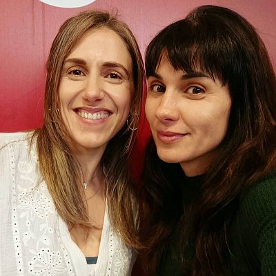 Primer plano de dos mujeres sonrientes muy juntas ante una pared roja, destacando la periodista Cristina Hermoso de Mena a la izquierda y otra mujer de cabello oscuro con flequillo a la derecha.