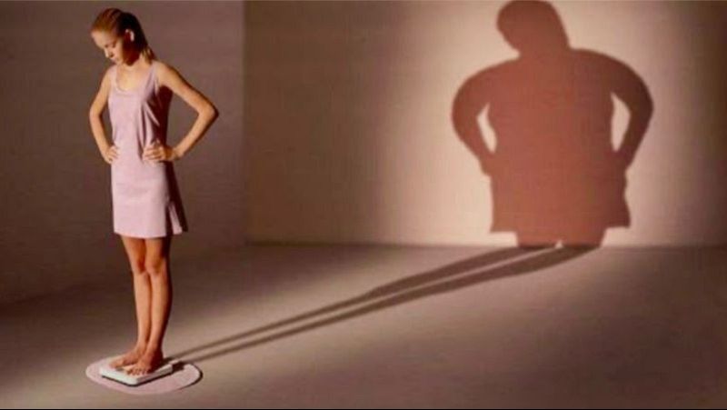 Mujer joven y delgada sobre una b�scula cuya sombra en la pared muestra una silueta con sobrepeso, ilustrando la dismorfia corporal en un ambiente de estudio minimalista.