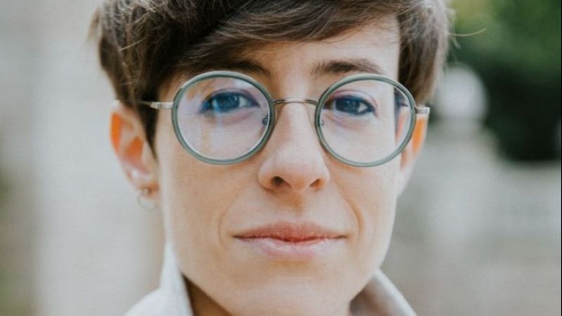 Primer plano frontal de la directora Irene Delgado con pelo corto, gafas redondas y una leve sonrisa, destacando sobre un fondo natural desenfocado.