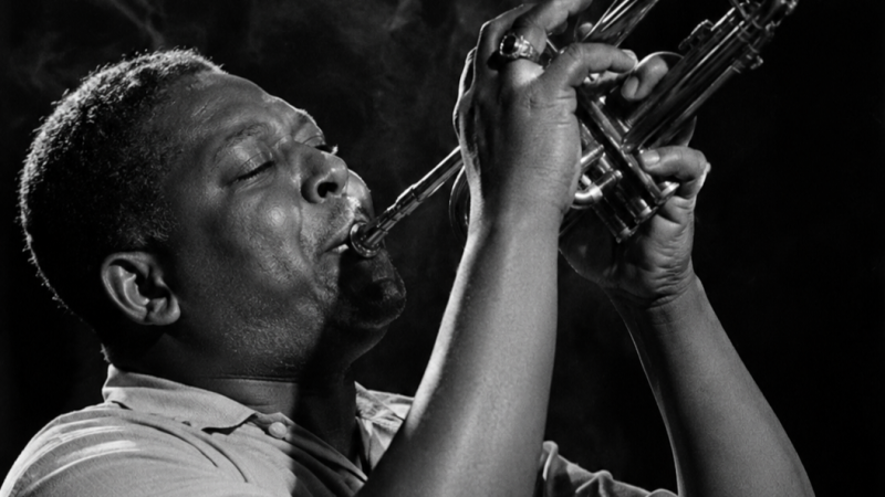 Primer plano en blanco y negro de un trompetista de jazz tocando con intensidad, ojos cerrados y rodeado de humo.