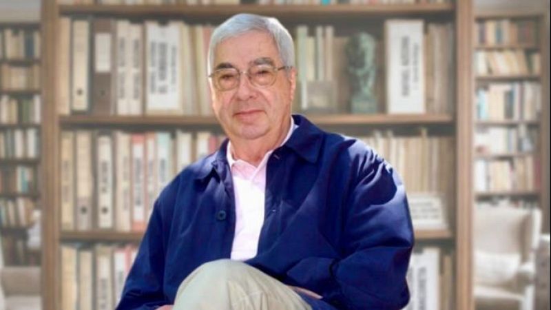 El escritor Rafael Azcona posa sentado en un plano medio corto frente a una estanter�a llena de libros y archivadores, luciendo chaqueta oscura y camisa clara.