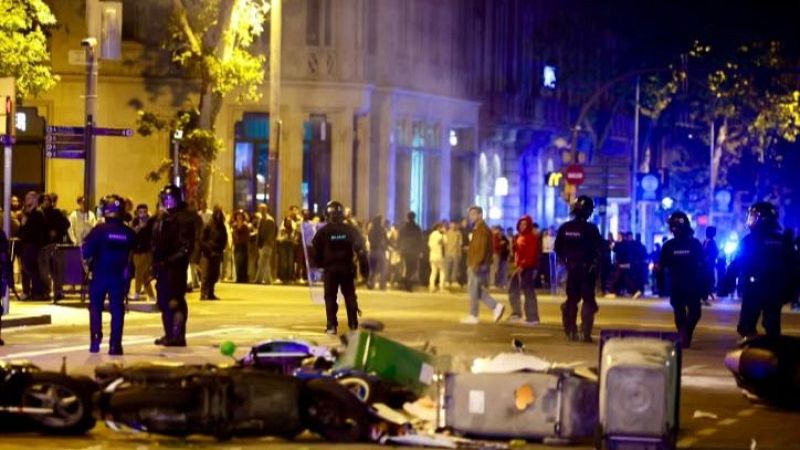 Calle urbana de noche con barricadas, contenedores volcados y agentes antidisturbios patrullando la calzada.