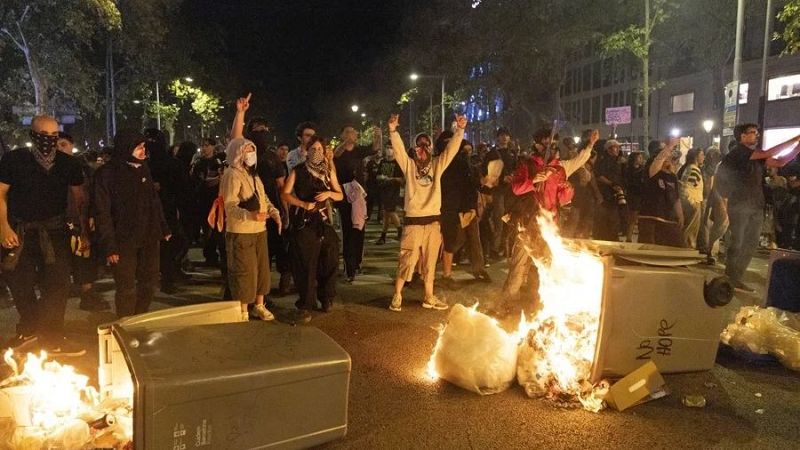 Contenedores de basura incendiados y manifestantes con los rostros cubiertos durante disturbios nocturnos en una calle de Barcelona.