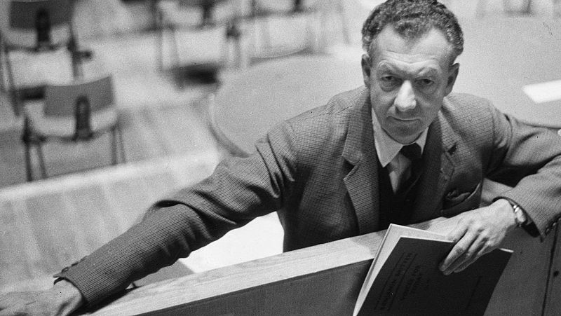 Retrato en blanco y negro del compositor Benjamin Britten apoyado en una barandilla de madera, sosteniendo una partitura con expresi�n seria.