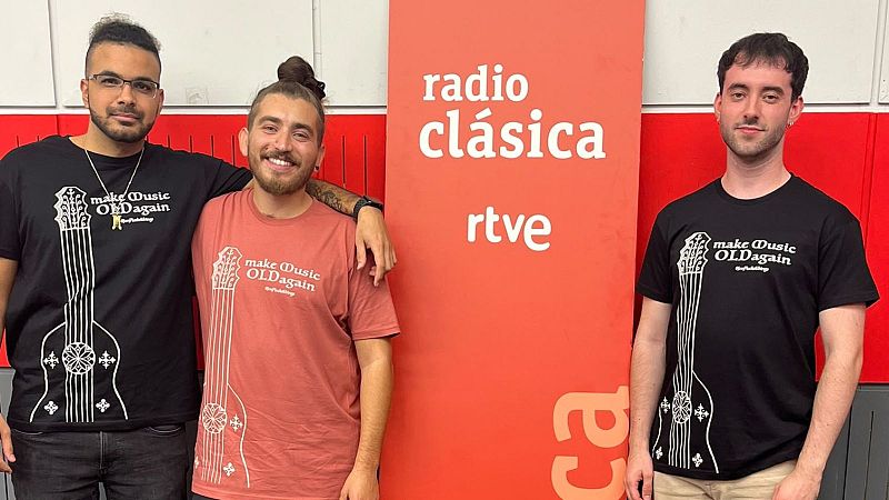 Tres j�venes sonrientes posan en un estudio de radio frente a un panel rojo, vistiendo camisetas con un dise�o de instrumento antiguo.