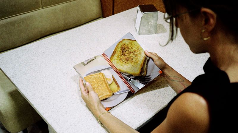 Mujer de espaldas con gafas y top negro hojea un libro abierto con fotos de sándwiches en una mesa de cafetería.