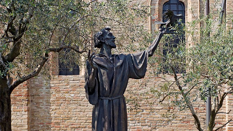 Escultura de bronce de San Francisco de As�s sosteniendo un p�jaro con el brazo extendido ante una pared de ladrillo r�stico.