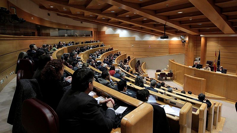 Sala de conferencias moderna con diseño semicircular en madera clara, asistentes en pupitres con micrófonos y mesa presidencial con banderas.