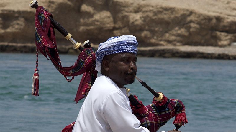 Un hombre con t�nica blanca y turbante oman� toca una gaita tradicional con fuelle de tart�n frente al mar y un acantilado des�rtico.