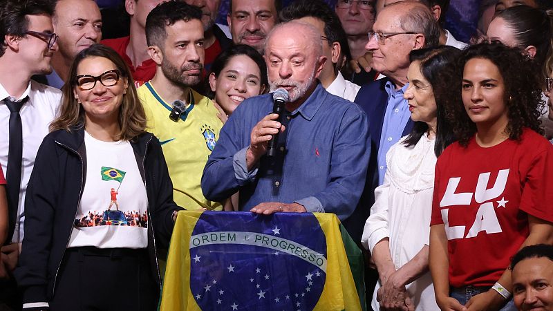Luiz Inácio Lula da Silva pronuncia un discurso tras su victoria electoral junto a su esposa y colaboradores, con la bandera de Brasil de fondo.