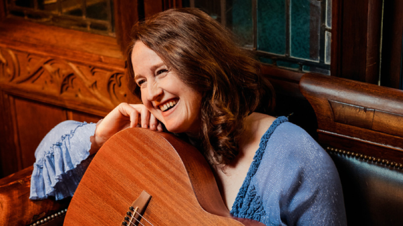 Plano medio de la cantautora Kate Rusby sonriendo alegremente, vistiendo blusa azul y abrazando una guitarra ac�stica en un interior cl�sico.