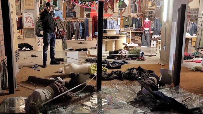 Interior de una tienda vandalizada con cristales rotos y maniqu�es ca�dos, mientras un hombre habla por tel�fono observando los destrozos.