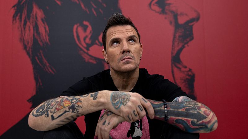Dani Mart�n, en imagen promocional de su disco, sentado con los brazos cruzados y tatuajes visibles, mira hacia arriba. Fondo rojo con su rostro ampliado.