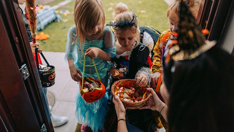 Un grupo de niños disfrazados, incluyendo una princesa, una pirata y un tigre, participan en la celebración de Halloween, recibiendo dulces de una persona adulta. En primer plano, una niña vestida de bruja sostiene un cubo decorado con motivos de Halloween.