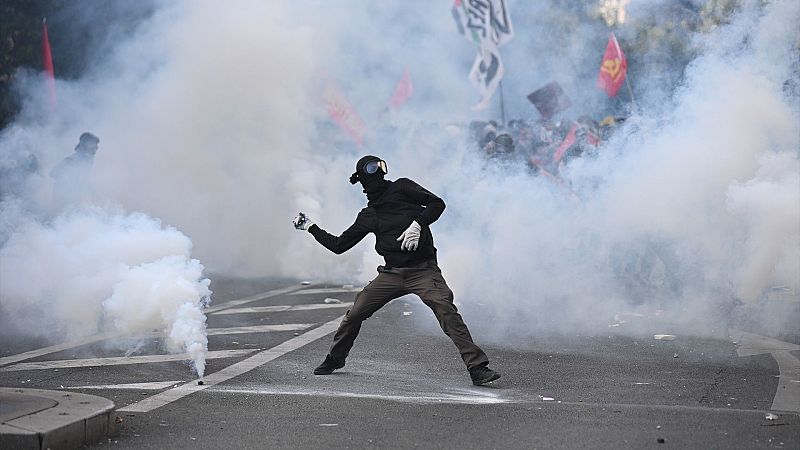 Una persona encapuchada y con gafas protectoras se sitúa en una calle con humo blanco, adoptando una postura de lanzamiento junto a manifestantes con banderas.