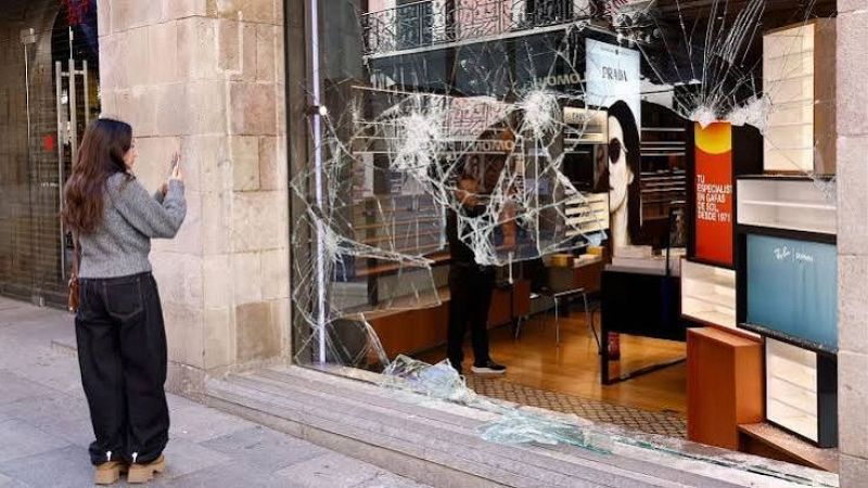 Escaparate destrozado de una �ptica con cristales rotos en la acera, fotografiado por una mujer de espaldas mientras se observa el interior con carteles publicitarios.