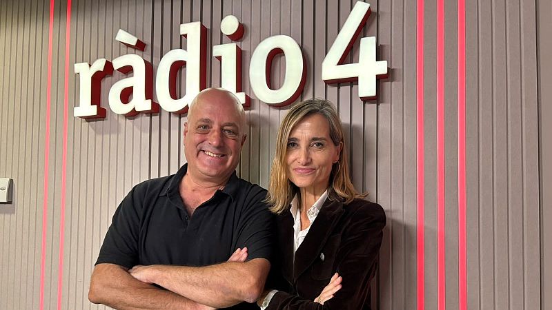Magda Carlas y un hombre posan sonrientes de pie y con los brazos cruzados en un estudio de radio con paneles de madera y luces rojas.