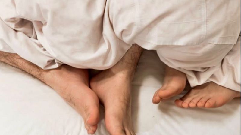 Primer plano de los pies descalzos de dos personas asomando bajo una s�bana blanca en una cama con iluminaci�n c�lida.