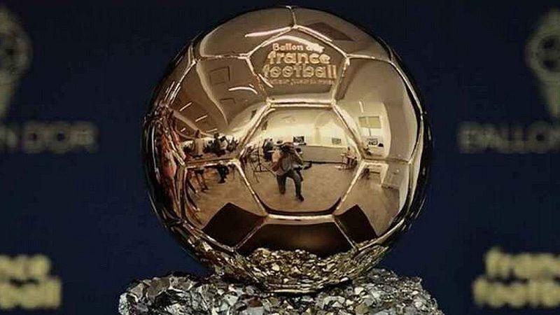 Primer plano del trofeo del Bal�n de Oro con esfera dorada reflectante sobre base de roca, mostrando el reflejo de un fot�grafo y fondo azul con texto corporativo.