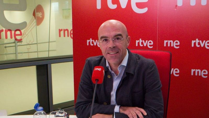 José Manuel Soria sonríe sentado ante un micrófono de radio en un estudio con panel acústico rojo y cristalera.