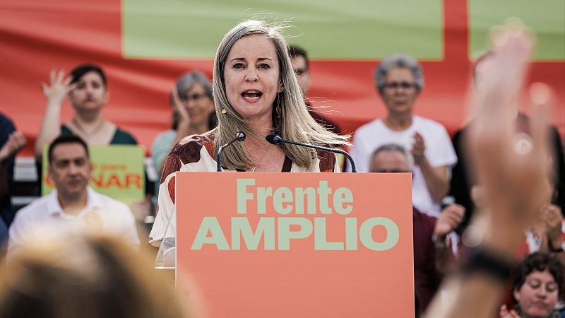 Plano medio de la política Verónica Barbero hablando ante un atril naranja con micrófonos en un mitin político al aire libre.