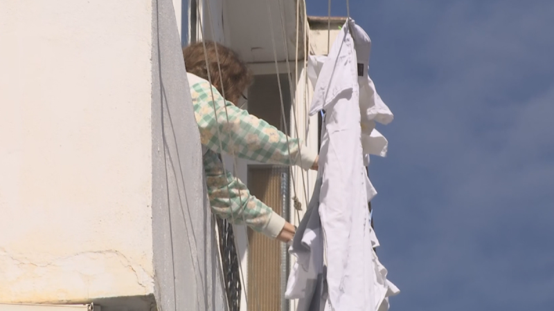 Una persona con gafas y jersey de cuadros tiende ropa blanca en un tendedero exterior bajo un cielo azul.