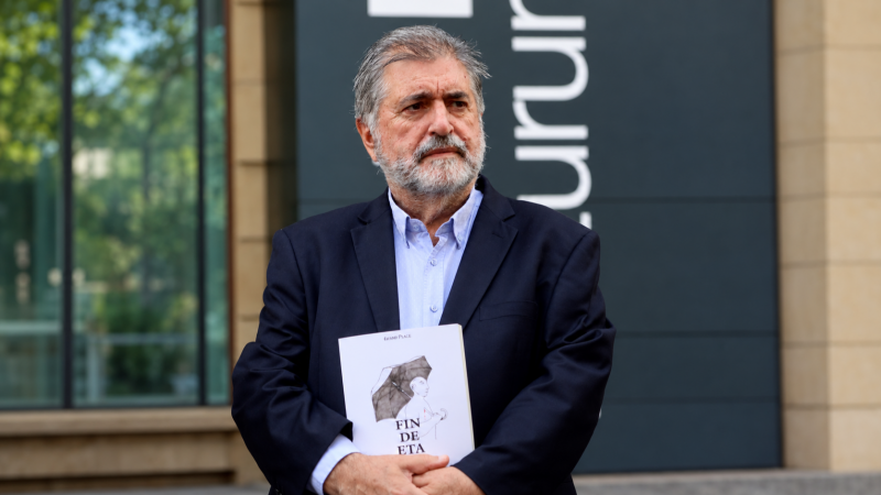 Plano medio de Jesús Eguiguren con americana azul marino, sosteniendo un libro titulado «FIN DE ETA» frente a un edificio moderno.