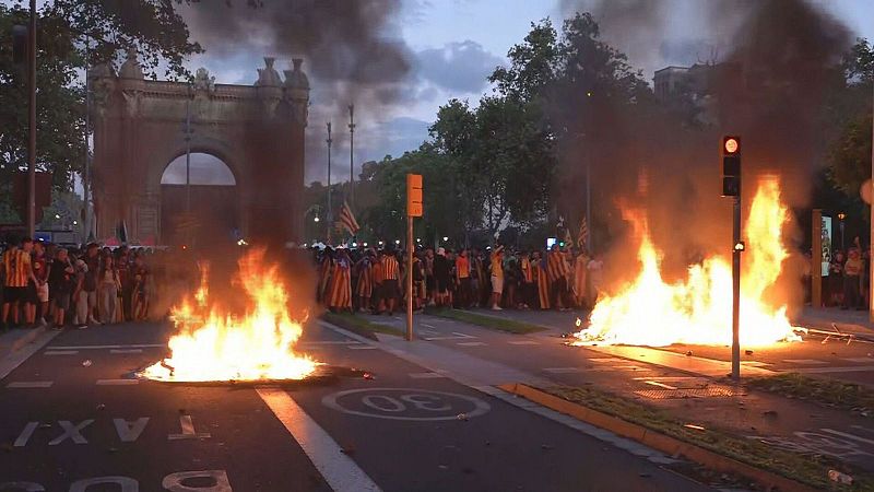 Barricadas en llamas cortan una amplia avenida urbana durante unos disturbios al anochecer, con manifestantes y el Arco de Triunfo al fondo.