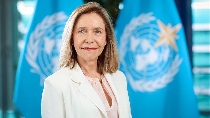 Retrato de Celeste Saulo con americana blanca y blusa rosa, sonriendo ante un fondo con banderas institucionales.