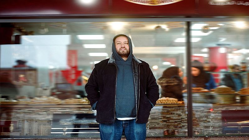 Hombre con barba y sudadera con capucha de pie ante un escaparate iluminado con reposter�a �rabe.