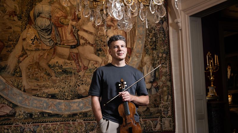 El violinista Johannes Pramsohler posa en una sala cl�sica junto a un tapiz antiguo, sosteniendo un viol�n y un arco bajo una l�mpara de ara�a.